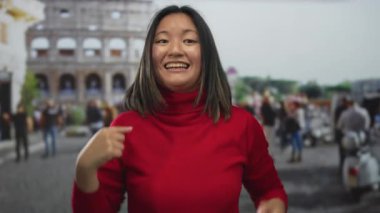 Kırmızı kazaklı Çinli kadın Roma Kolezyumu 'nda neşeli bir ifadeyle kutlama yapıyor. Şehir kalabalığının arasında canlı bir şehir ortamını vurguluyor..