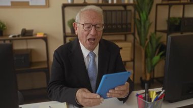 Takım elbiseli kıdemli işadamı tabletle video görüşmesi yapıyor. Modern ofis ortamında gülümsüyor. Bitkiler ve ofis malzemeleriyle profesyonel ama sıcak bir ortam yaratıyor..