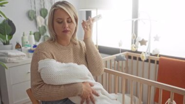 Kadın yatak odasındaki beşikte kundaklanmış bebeği nazikçe kulağına tutarken tutuyor; annelik sorumluluğu..