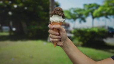 Güneşli bir günde parkta elinde çikolatalı ve vanilyalı dondurma külahı olan bir adam yeşil ağaçlarla çevrili yaz tatilinin tadını çıkarıyor.