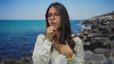 Sahilde gözleri kapalı ve eli göğsünde öksüren, gözlüklü, etrafı okyanus ve taşlarla çevrili bir kadın, gün ışığında dışarıda dururken sağlık endişesi gösteriyor..