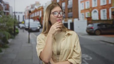 Gözlük takan kadın gündüz vakti şehir sokağında dikilirken çenesine hafifçe dokunur..