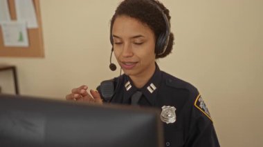 Polis üniformalı bir kadın ofis ortamında masasında kulaklık takıyor, profesyonel çevrede ekipman ve belgelerle çalışırken odaklanıyor ve özen gösteriyor.