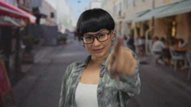 Kalabalık bir şehir caddesinde parmakla gösteren ve gülümseyen bir kadın elleri olan bir kalp yaratıyor, arka planda insanların yemek yediği bir şehir ortamında neşe sergiliyor..