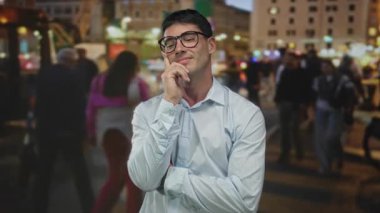 Gün batımında kalabalık bir sokakta el pençe üzerinde duran gözlüklü ve açık renk gömlekli bir adam şehrin ışıkları altında düşünüyor..