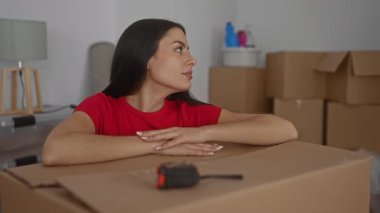 Yeni apartman dairesinde kırmızı gömlekli bir kadın büyük bir karton kutuya gülümseyen ve yaslanan kutularla çevrili, yeni bir eve taşınmanın sevincini ve beklentisini yakalıyor..