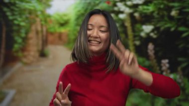 Kırmızı kazaklı genç bir kadın açık hava parkında gülümsüyor ve barış işareti gösteriyor.