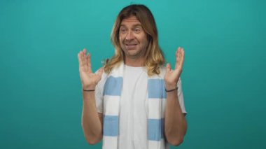 Çizgili futbol eşarbı takan sarışın adam, stüdyoda tezahürat jestinde elini kaldırıyor; takım desteği coşkusu.
