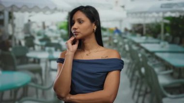 Teras restoranında dikilen bir kadın, düşünceli bir ifadeyle sahne düşünüyor, arka planda masalar ve sandalyeler, mavi elbise, parlak gündüz atmosferi..