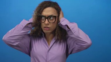 Transseksüel kadın, stüdyoda, mor gömlek ve gözlük takarak kulaklarını mavi arka plana kapatır; kimlik dayanıklılığı endişesi.