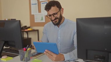 Modern ofis ortamında tablet kullanan, bilgisayar ve kırtasiyeyle çevrili İspanyol sakallı bir adam, kapalı alanda verimli bir çalışma alanı sergiliyor..