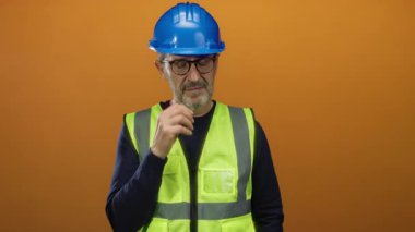 Mavi kasklı ve yansıtıcı yelek giyen mühendis parlak turuncu bir arka plana karşı düşünceli bir şekilde duruyor..