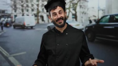 Siyah üniformalı İspanyol şef, kalabalık bir caddede durup, yoldan geçenlere gülerek enerjik bir şekilde el hareketi yapar..