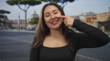 Uzun kahverengi saçlı ve siyah bluzlu bir kadın sokakta parmaklarını yanağına uzatırken gülümsüyor; mutluluk..