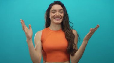 Turuncu bluz giyen ve gülümseyen genç bir kadın stüdyoda turkuaz arka planda kollarını açıyor; mutluluk..