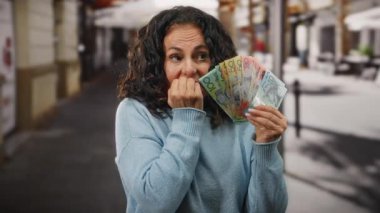 Bir kadın endişeyle bir şehir caddesinde elinde banknotlarla mavi bir kazak giyiyor, mali kaygıları ve duyguları temsil ediyor..