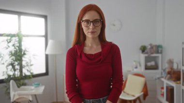 Kızıl saçlı bir kadın, rahat bir yatak odasında gülümsüyor, gözlük takıyor ve kırmızı bir kazak giyiyor, arka planda lamba ve bebek eşyalarıyla evde duruyor, sıcaklık ve cazibe saçıyor..