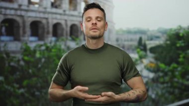 Roma Kolezyumu 'nun yakınında meditasyon yapan genç bir adam, sakin bir ifade sergiliyor, arka planda yemyeşil bir ortam var, kentsel bir ortamda sükunet öneriyor..