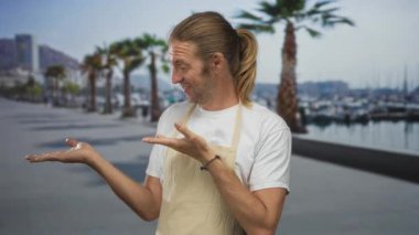 Önlük takan uzun saçlı sarışın adam geniş bir gülümseme takınıyor ve sokakta avuçlarını açıyor; neşe.