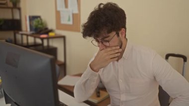 Ofis odasındaki bilgisayarda çalışan sakallı genç adam düşünceli ve meşgul görünüyor. Etrafı kitaplar ve bitkilerle çevrili. İçerde sıradan ama profesyonel bir ortam yaratıyor..