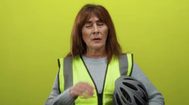 Sarı yelek giymiş İspanyol kıdemli bir kadın elinde bir kask tutarken boynunu parlak sarı bir zemin üzerinde tutarak rahatsızlık ve hayal kırıklığını ifade ediyor..