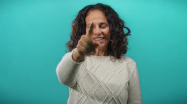 Kıvırcık saçlarıyla kameraya doğru gülümseyen, parlak mavi bir arka plana bakan, rahat beyaz bir kazak giyen ve neşeli ve çekici bir ruh halini canlandıran bir kadın..