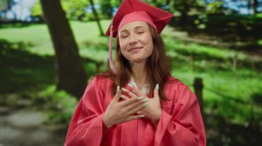 Mezuniyet elbisesi giymiş, elleri yüreğinde güneşli bir park ortamında gülümseyen, başarısını ve neşesini ön plana çıkaran genç bir kadın..