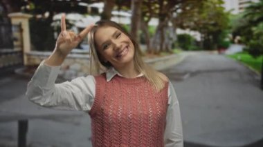 Arka planda ağaçlar olan bir şehir caddesinde taştan bir jest yapan genç bir kadın, açık hava kentsel ortamında güven ve neşe ifade ediyor..