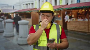 Şehir pazarında elinde para tutan adam, güçlü bir kokuya tepki olarak burnunu kapatıyor. Sarı bir şapka ve yansıtıcı yelek de dahil olmak üzere güvenlik kıyafeti giyiyor..