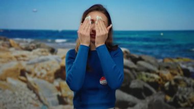 Plajda oy kartı ve mavi kazak giyen genç bir kadın deniz manzaralı bir arka planda elleri olan neşeli yüzler çiziyor..