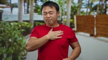 Açık havada göğsünü tutan kırmızı gömlekli genç adam bulanık şehir geçmişiyle çevrili rahatsızlığını dile getiriyor. Açık hava ortamında kalp ağrısı ya da sağlık sorunu olduğunu gösteriyor..