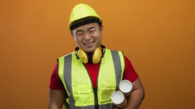 Elinde planlar olan genç adam turuncu bir arka plana karşı güvenlik yeleği ve kask giyiyor. İnşaat projelerine hazır kendine güvenen bir mimar ya da mühendisi resmediyor..