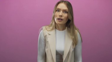 Pembe izole edilmiş arka planda el kol hareketi yapan, sarışın ve bej elbiseli, stüdyo ortamında çeşitli duygular gösteren bir kadın..