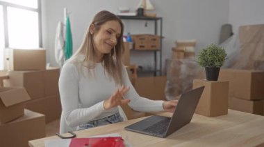 Yeni evdeki kadın video konferansı modern oturma odasındaki dizüstü bilgisayarla kutuları taşıyor..