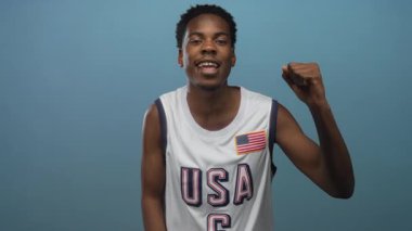 Amerikan bayraklı basketbol forması giyen adam gülümseyip soluk mavi stüdyoda elini kaldırıyor..