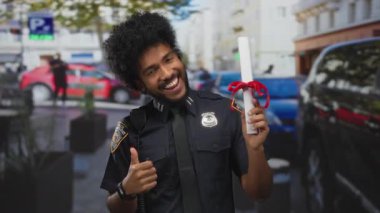 Polis üniformalı kendinden emin bir adam. Sokak ortamında diplomasıyla gülümsüyor. Açık havada başarılar sergiliyor..