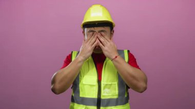 Sarı şapka ve güvenlik yeleği giyen bir adam pembe arka planda gözlerini ovuşturuyor. Bu da yorgunluk ve inşaat yorgunluğunu gösteriyor..