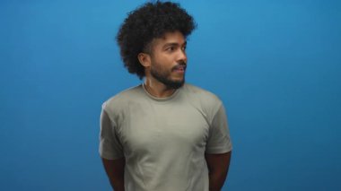 Kıvırcık saçlı, gülümseyen ve parlak mavi bir arka plana işaret eden bir adam güven ve neşe saçar.