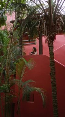 Zengin palmiyeler ve canlı mimari ile çevrili tropikal bir tatil beldesinde balkon manzarasından hoşlanan bir kadın, egzotik bir otel ortamında lüks ve rahatlığı temsil ediyor.