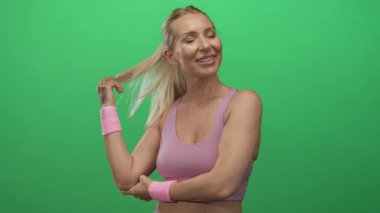 Sarışın kadın yeşil stüdyoda pembe bilekliğin üzerinde dinlenirken gülüyor ve at kuyruğu sallıyor; tefekkür.