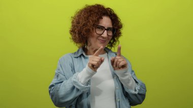 Orta yaşlı, esmer bir kadın canlı sarı arka plana karşı öpücük hareketi yapıyor. Günlük kıyafetler ve gözlük takıyor. Neşeli duygular ifade ediyor..