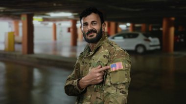 Askeri üniformalı İspanyol adam park yerinde duruyor, kolundaki Amerikan bayrağını gösteriyor, etrafı park halindeki arabalarla çevrili ve loş bir garajla çevrili..