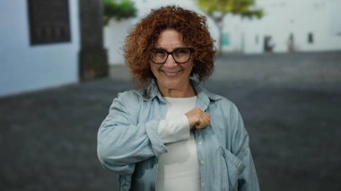 Kıvırcık saçlı kadın gülümsüyor ve şehir sokaklarındaki bir kameraya yumruğunu gösteriyor. Açık hava ortamında güç ve kendine güveni somutlaştırıyor..