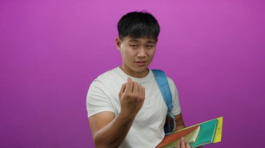 Genç Çinli erkek öğrenci, canlı pembe bir arkaplan üzerinde beyaz bir gömlek ve renkli klasörler tutarak işaret ediyor..