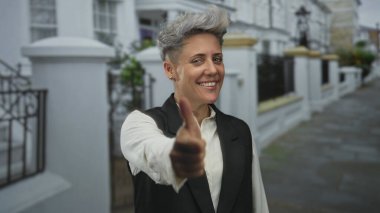 Beyaz teraslı binalarla dolu hareketli caddede gülümseyen ve başparmak kaldıran bir kadın. Onaylama pozitifliği..