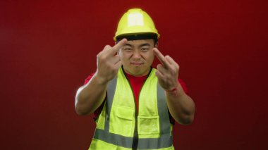 İnşaat işçisi koyu kırmızı arka planda anlamlı bir jest yapıyor sarı kaskını ve yansıtıcı yeleğini vurguluyor..