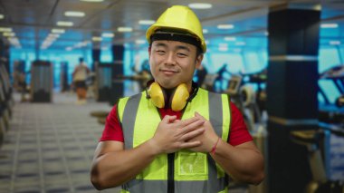 Spor salonundaki adam kasketli ve kulaklıklı, gülümseyen ve göğsünde el ele tutuşan parlak iç mekandaki egzersiz ekipmanlarının arasında minnettarlığını dile getiriyor..