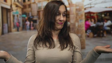 Koyu renk saçlı ve bej renkli jestleri olan bir kadın açık bir sokak terasında meraklı ve etkileşim gösteren bir restoran ortamında ellerini açık tutuyor..