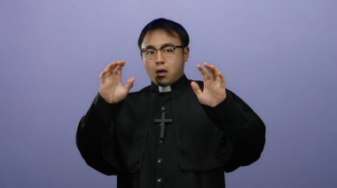 Papaz kıyafetli genç adam, izole bir mor arka planda, kaldırılmış eller ve kaldırılmış gözlüklerle, şok ve dini aktararak irkildi..