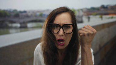 Gözlüklü ve uzun saçlı bir kadın eski bir kasaba caddesinde yumruğunu kaldırıyor canlı ve canlı bir şehir ortamında kararlılığını ifade ediyor..